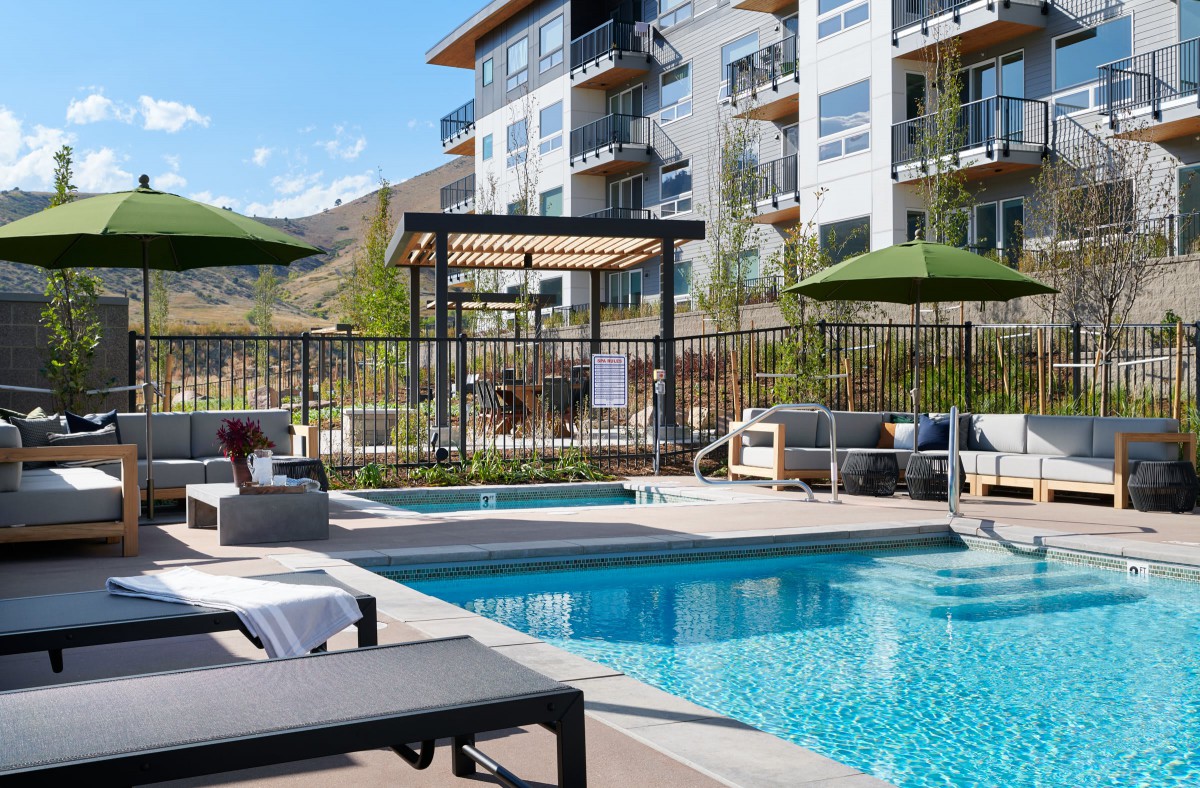 A modern apartment building features a luxurious outdoor swimming pool area with loungers and patio furniture on a sunny day.