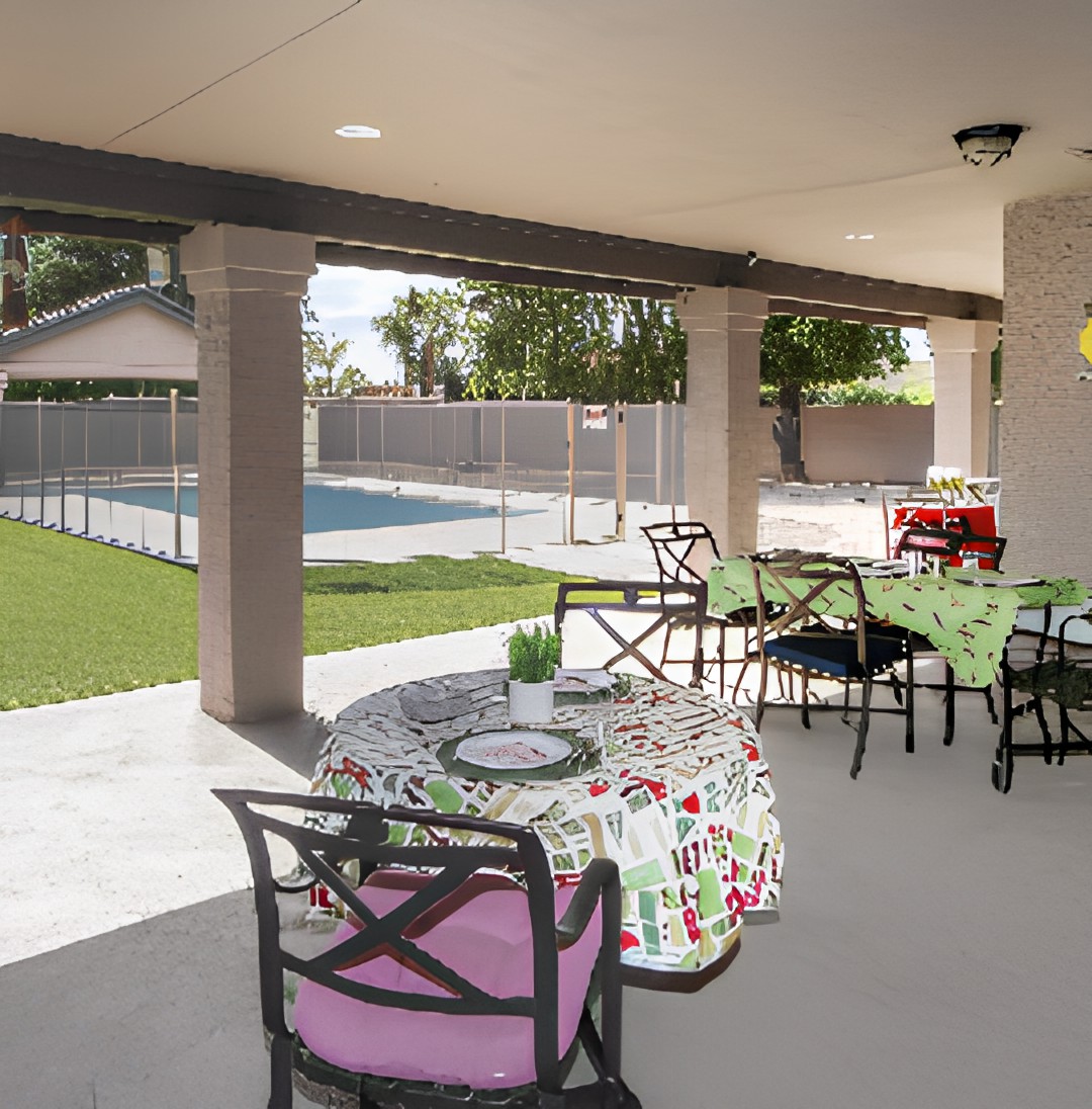 A covered patio with tables and chairs overlooking a garden and swimming pool.