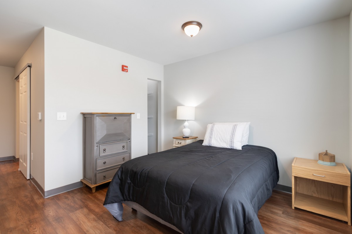 A minimalist bedroom with a bed, dresser, and lamp on a wooden floor.