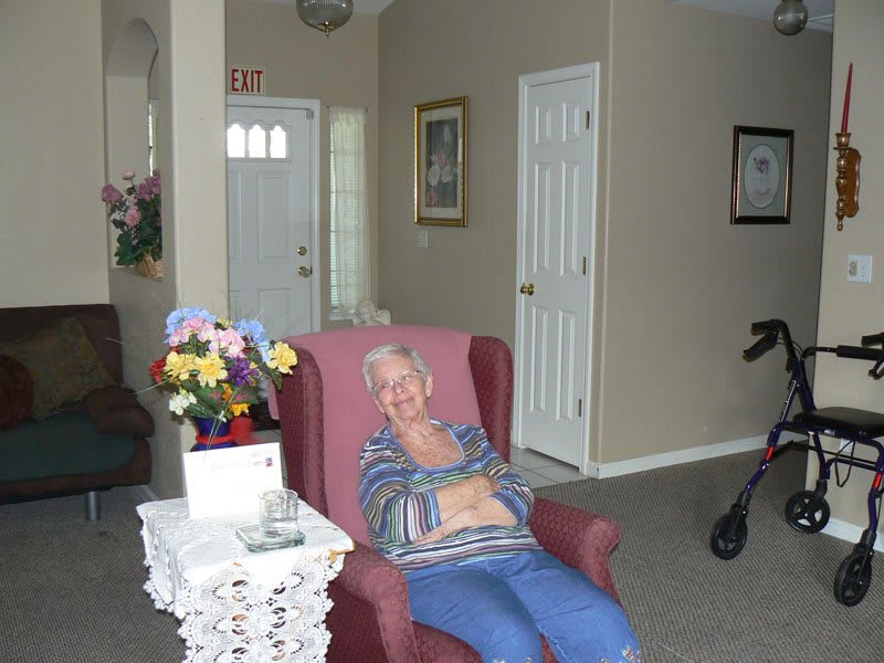 An elderly woman sitting comfortably in an armchair in a cozy living room with flowers and a walker nearby.