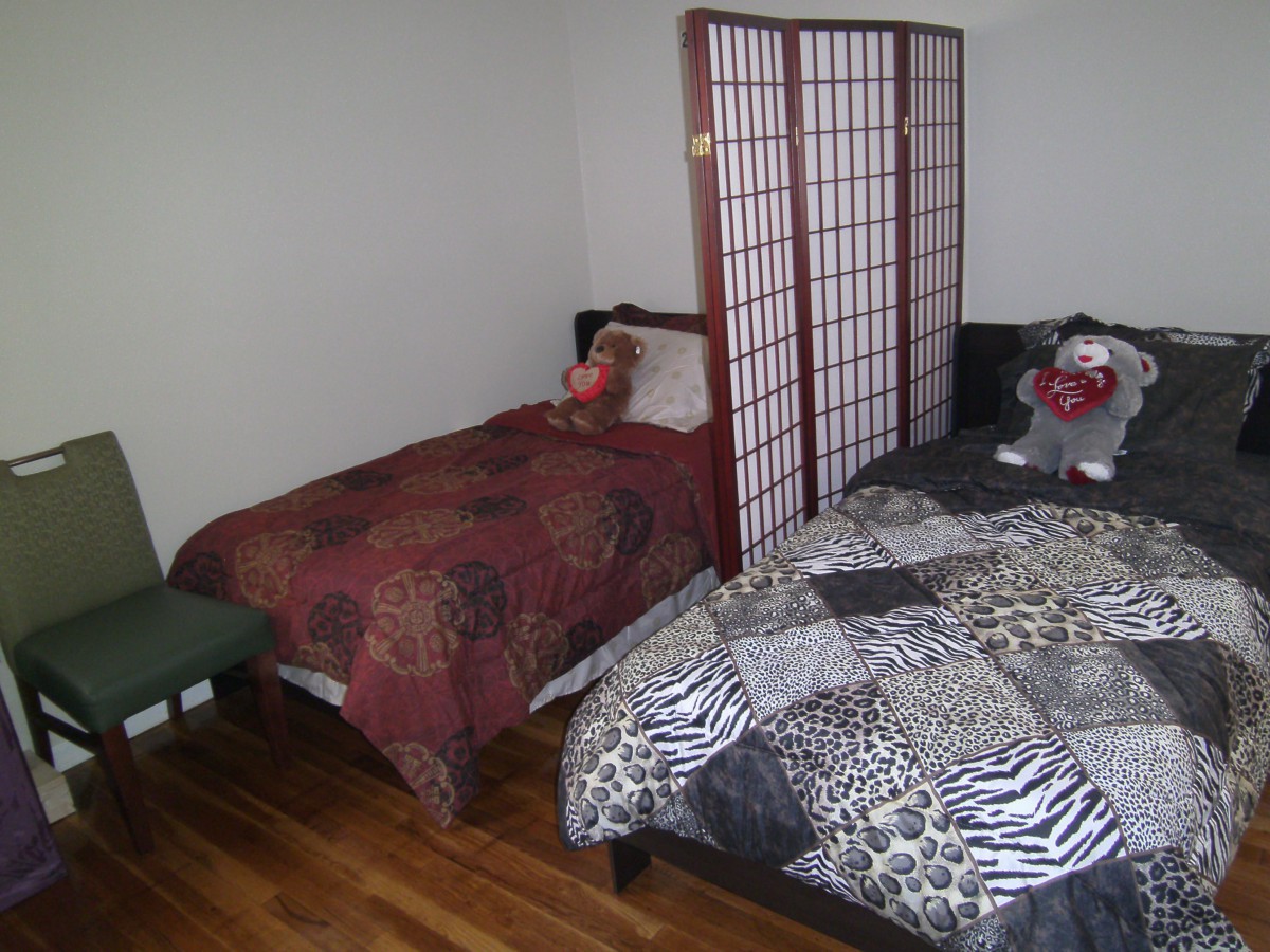 A cozy bedroom with two beds separated by a screen divider, featuring wooden floors and a teddy bear on one bed.