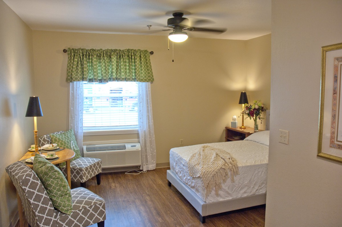 A cozy bedroom with a neatly made bed, two chairs, a small table, and a window adorned with green curtains.