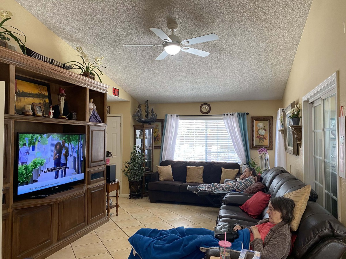 A cozy living room with a person relaxing on the couch while watching TV, featuring a ceiling fan and large window.