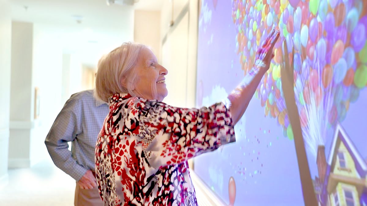 An elderly woman engaging with interactive art on a digital screen, smiling as she touches the colorful display.