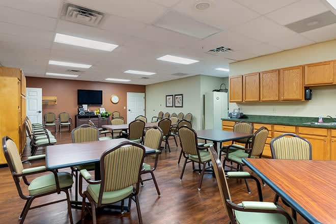 A spacious community room with multiple tables and chairs arranged for gatherings, featuring a kitchenette in the corner.
