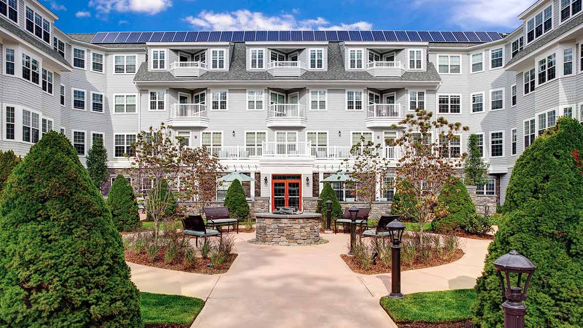 A modern apartment building with solar panels, featuring a landscaped garden and outdoor patio area.