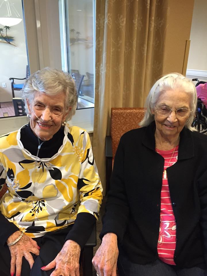 Two elderly women seated indoors, smiling at the camera.