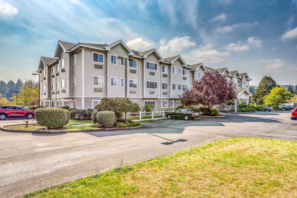 A exterior view of a multi-story apartment building with landscaped surroundings and a parking area on a sunny day.