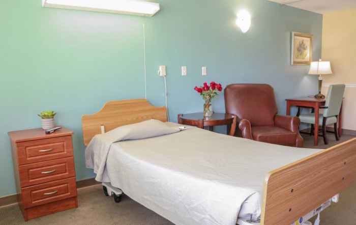 A hospital room with a bed, chair, and vase of roses.