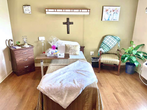 A cozy hospital room featuring a bed with a blanket, a small table with flowers, and a chair for visitors.