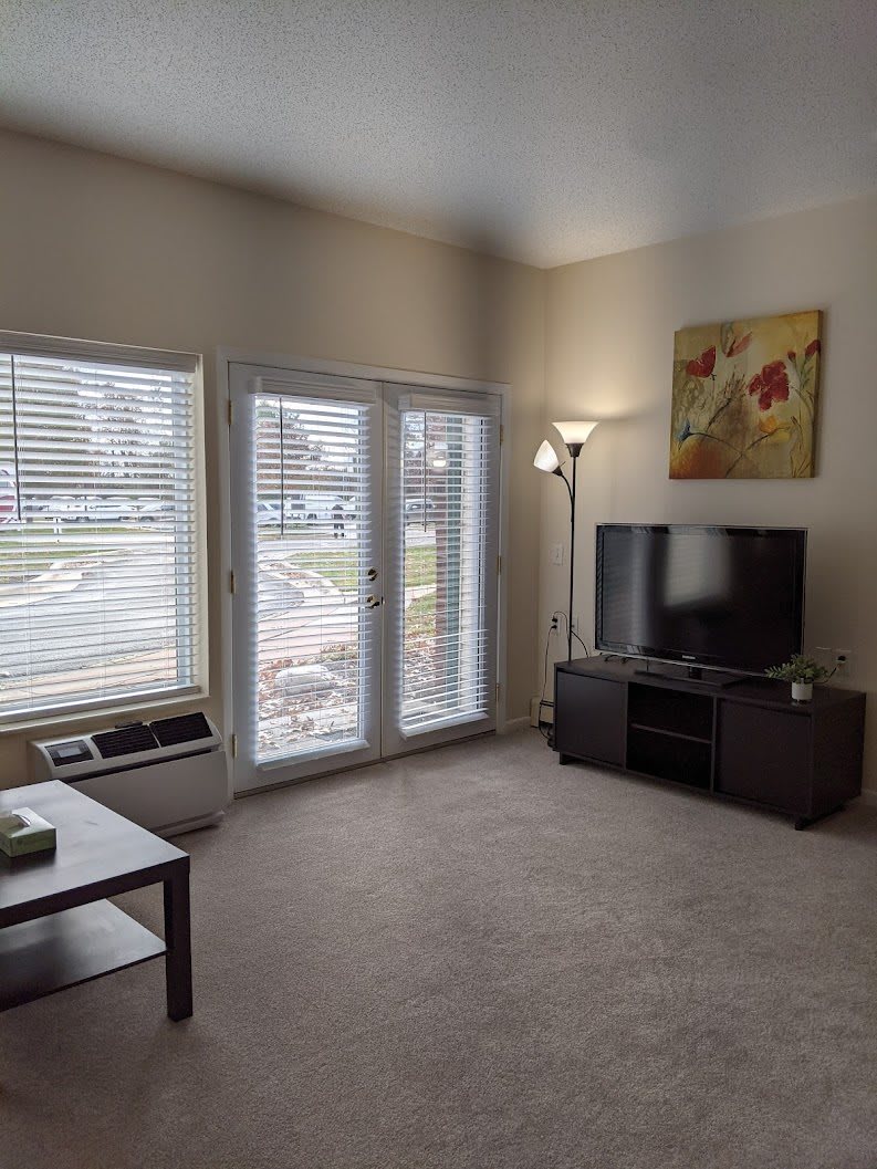 A cozy living room with a television, carpet, and large windows with blinds.