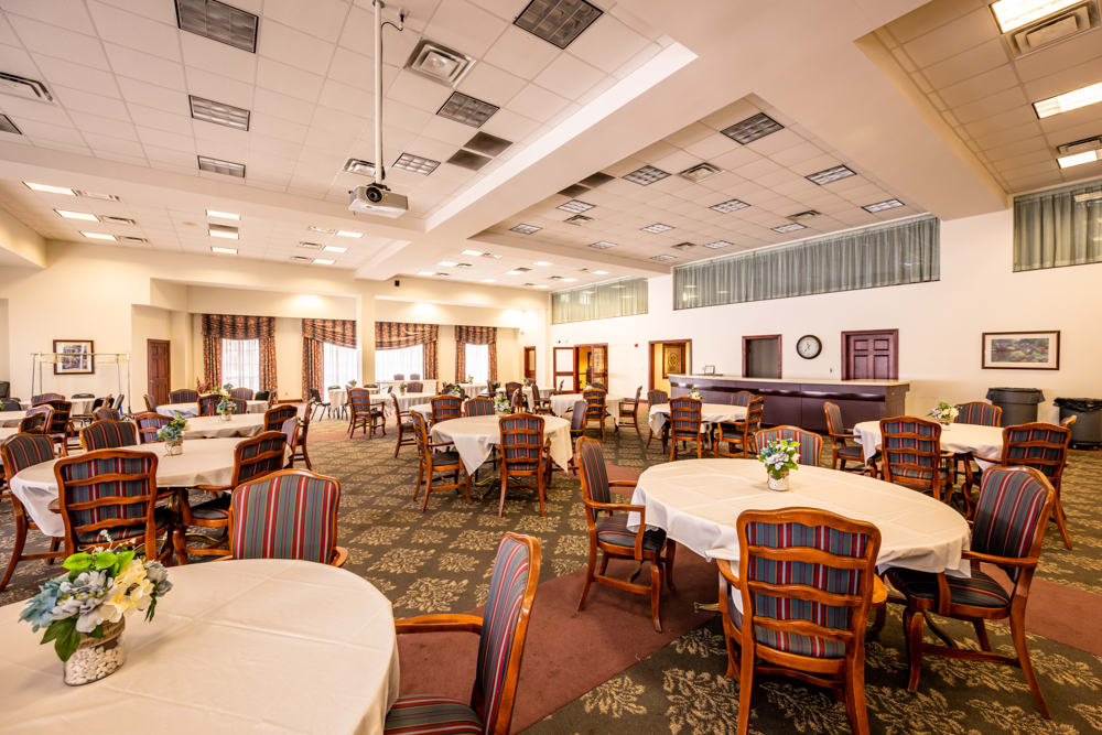 A spacious dining room with round tables and upholstered chairs, featuring warm lighting and carpeted flooring.