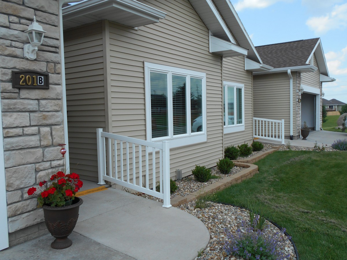 A house exterior with beige siding, white-trimmed windows, and a small porch with railing surrounded by neatly landscaped plants.