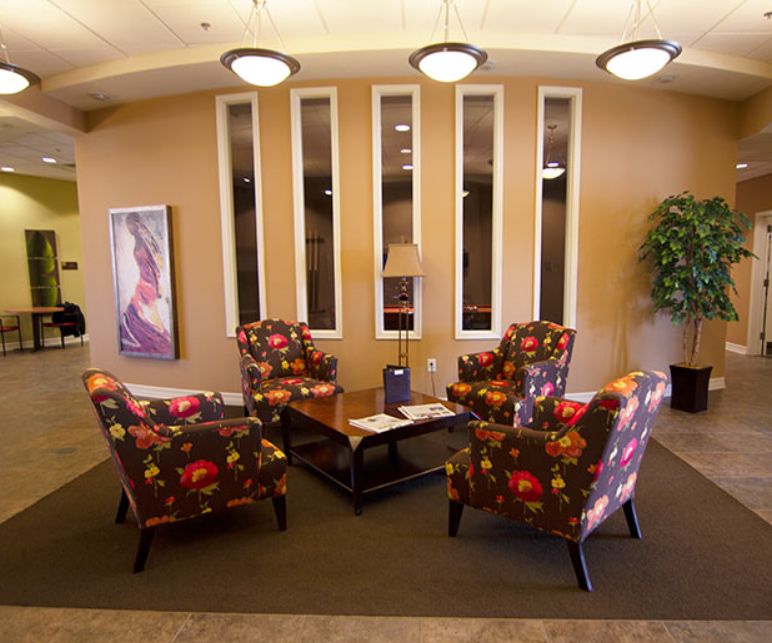 A cozy waiting room featuring floral armchairs, a coffee table, decorative plants, and wall art.