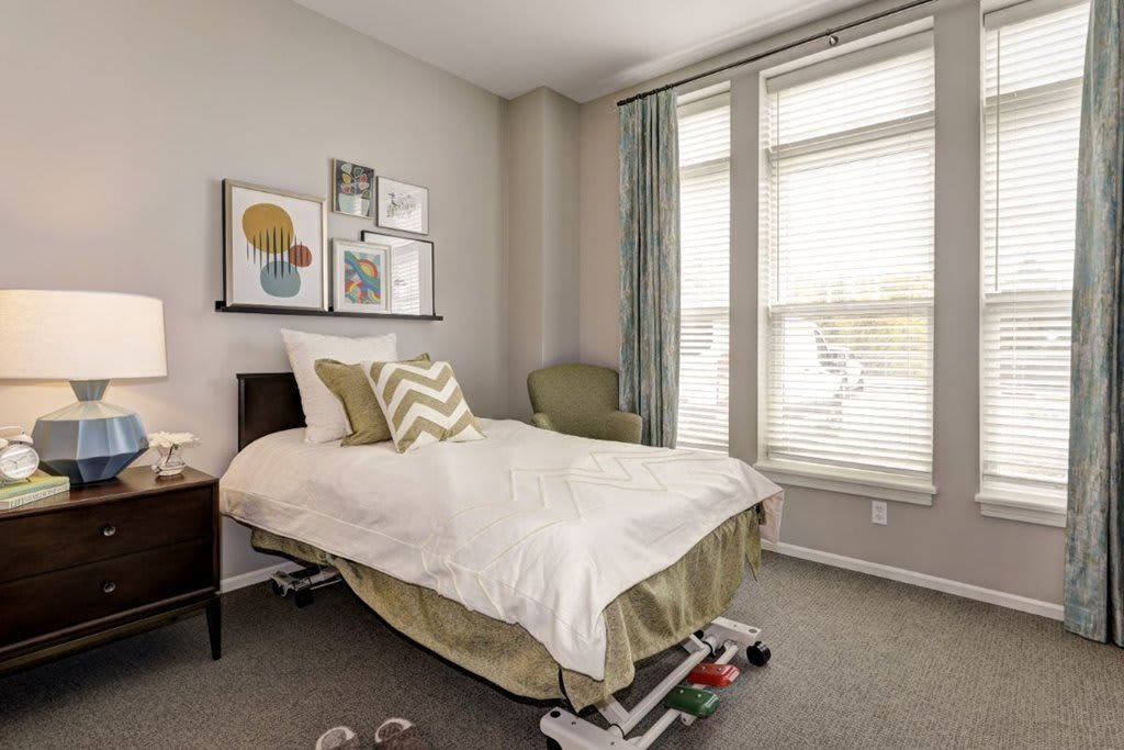 A cozy bedroom with a neatly made bed, artwork on the wall, and large windows.