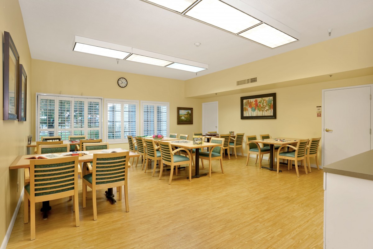 A spacious dining room with multiple tables and chairs, illuminated by overhead lighting and natural light from large windows.