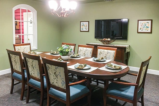 A formal dining room with a large table set for eight, illuminated by a chandelier and featuring a wall-mounted television.