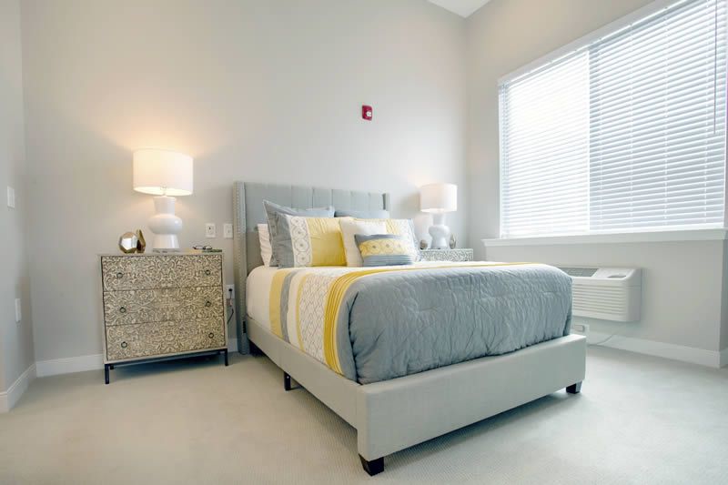 A modern and minimalist bedroom featuring a neatly made bed with gray and yellow bedding, flanked by two lamps on a wooden nightstand.
