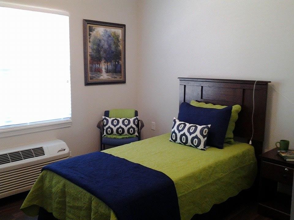 A modern bedroom with a single bed featuring green and blue bedding, complemented by a decorative pillow and artwork on the wall.