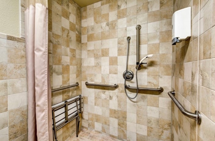 An accessible shower featuring tiled walls, grab bars, a folding seat, and a handheld showerhead.
