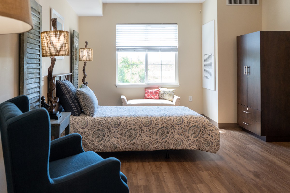 A cozy bedroom with ample natural light, featuring a bed, chair, and wooden flooring.