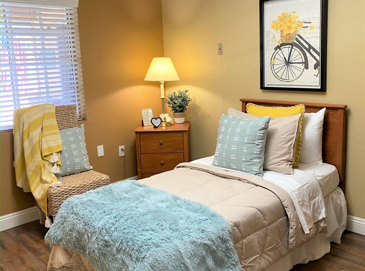 A cozy bedroom with a neatly made bed, featuring soft blue and beige bedding, decorative pillows, and warm lighting from a bedside lamp.
