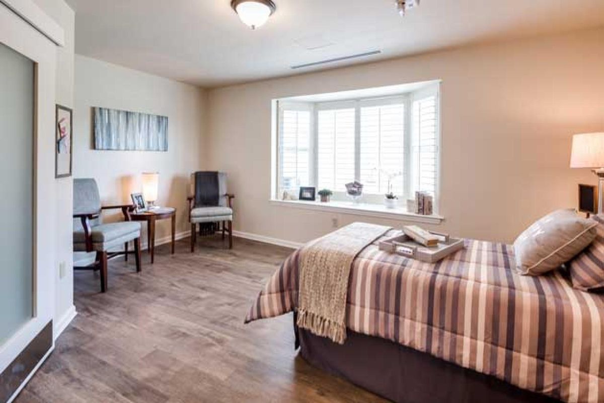 A cozy bedroom with a bed, armchair, and desk by a large bay window.