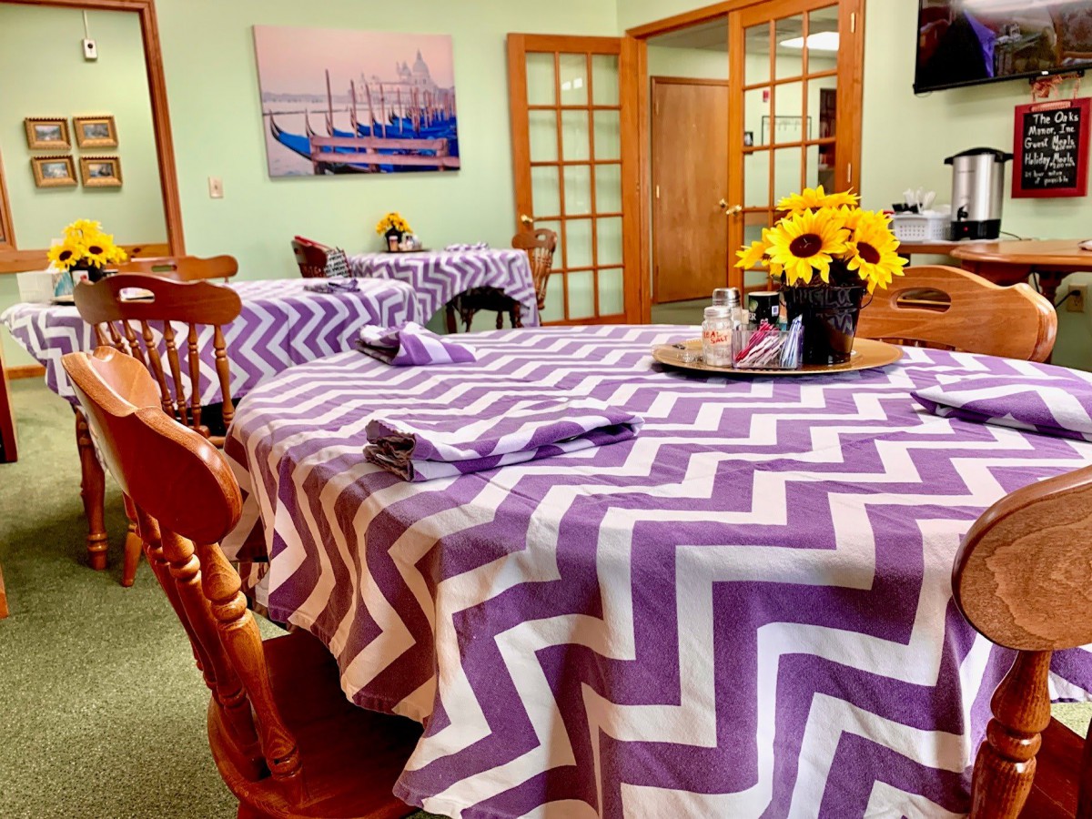 A cozy dining room with tables covered in purple zigzag patterned tablecloths, adorned with a vase of sunflowers.