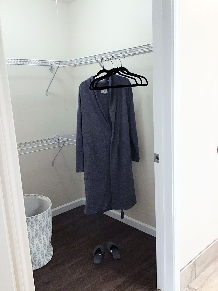An empty closet with a single robe hanging on black hangers, a laundry basket and shoes on the floor.