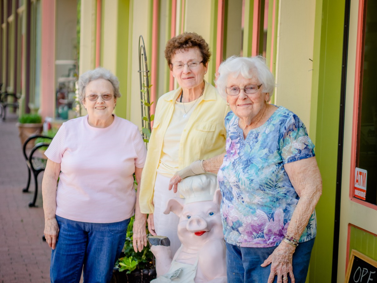 Three elderly women pose together outdoors with a small statue.