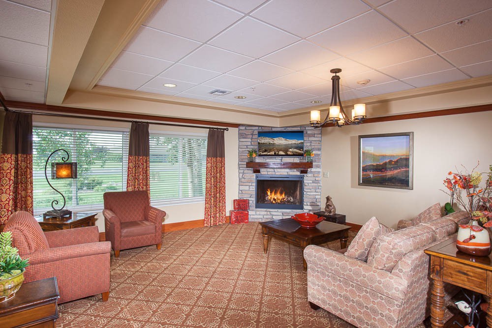 A cozy living room with a fireplace, sofa, and armchairs, complemented by a warm chandelier and large windows offering a scenic view.