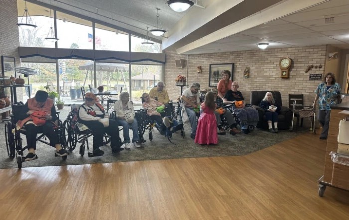 A group of seniors gathered in a nursing home common area, some seated in wheelchairs.