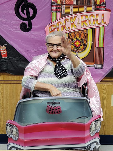 An elderly woman in a pink cape greets warmly while sitting in a toy car, embodying joy and vintage charm.