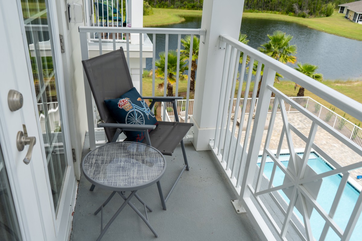 A cozy balcony setup with a chair, cushion, and table overlooking a lake and palm trees.