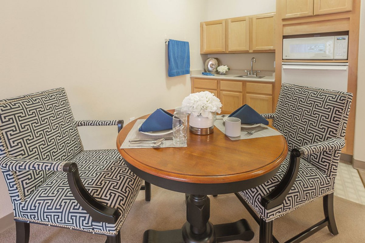 A cozy dining area featuring a wooden round table set for two, with patterned chairs and a minimalist kitchen backdrop.