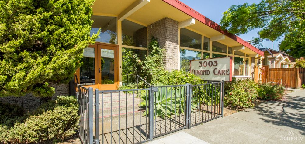 A modern building entrance with large glass windows, greenery, and a sign reading '3005' surrounded by a metal fence.