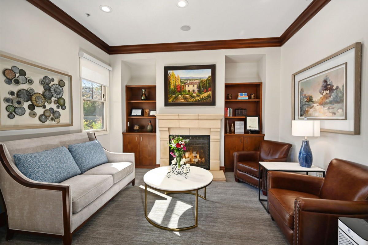 A cozy and elegant living room with a fireplace, featuring a sofa, armchair, coffee table, bookshelves, and artwork.