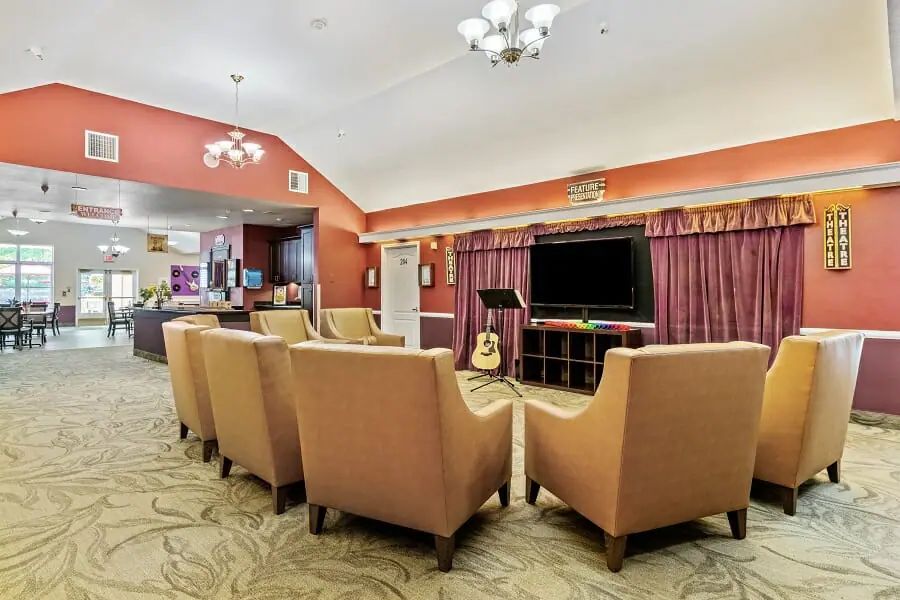 A cozy living room setup with several chairs facing a television, a guitar leaning against the TV stand, and elegant chandeliers hanging from the ceiling.