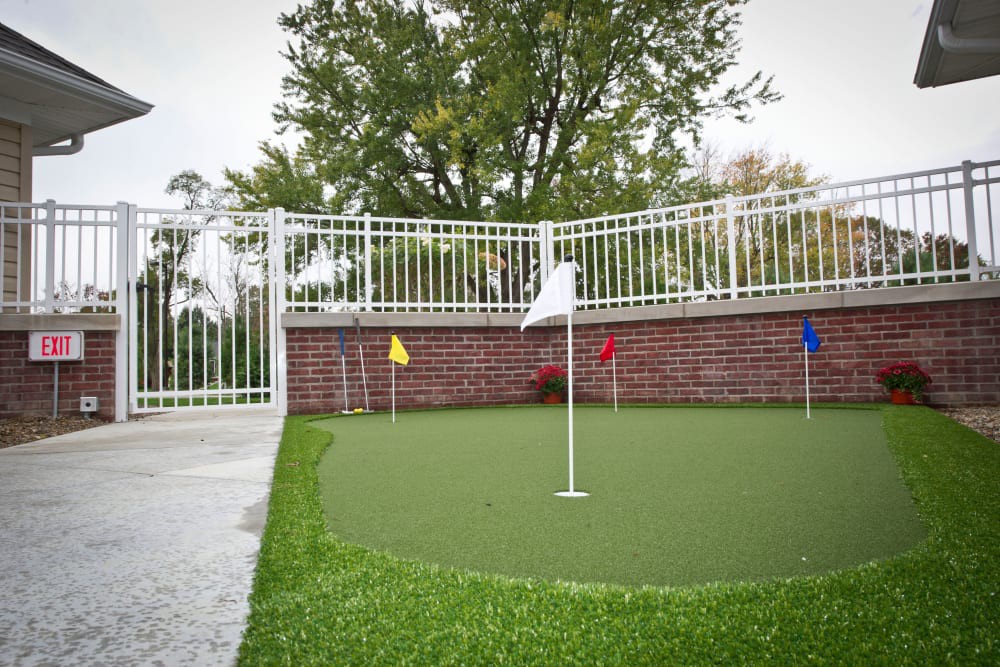 An outdoor putting green with multiple colored flags, set against a brick wall and fence.
