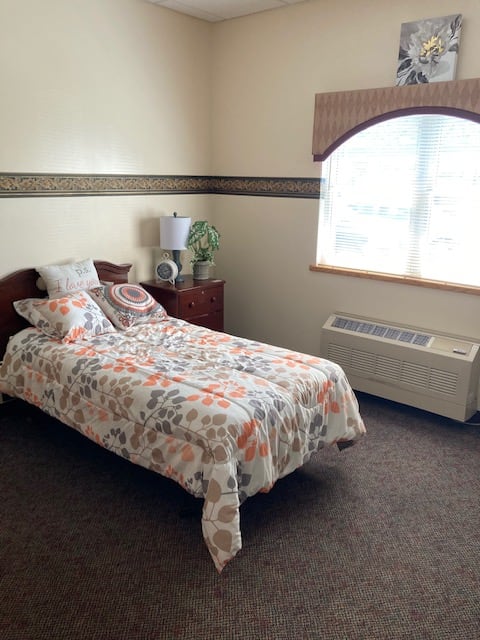 A cozy bedroom with a single bed featuring floral bedding, a wooden nightstand with a lamp, and a large window allowing natural light in.