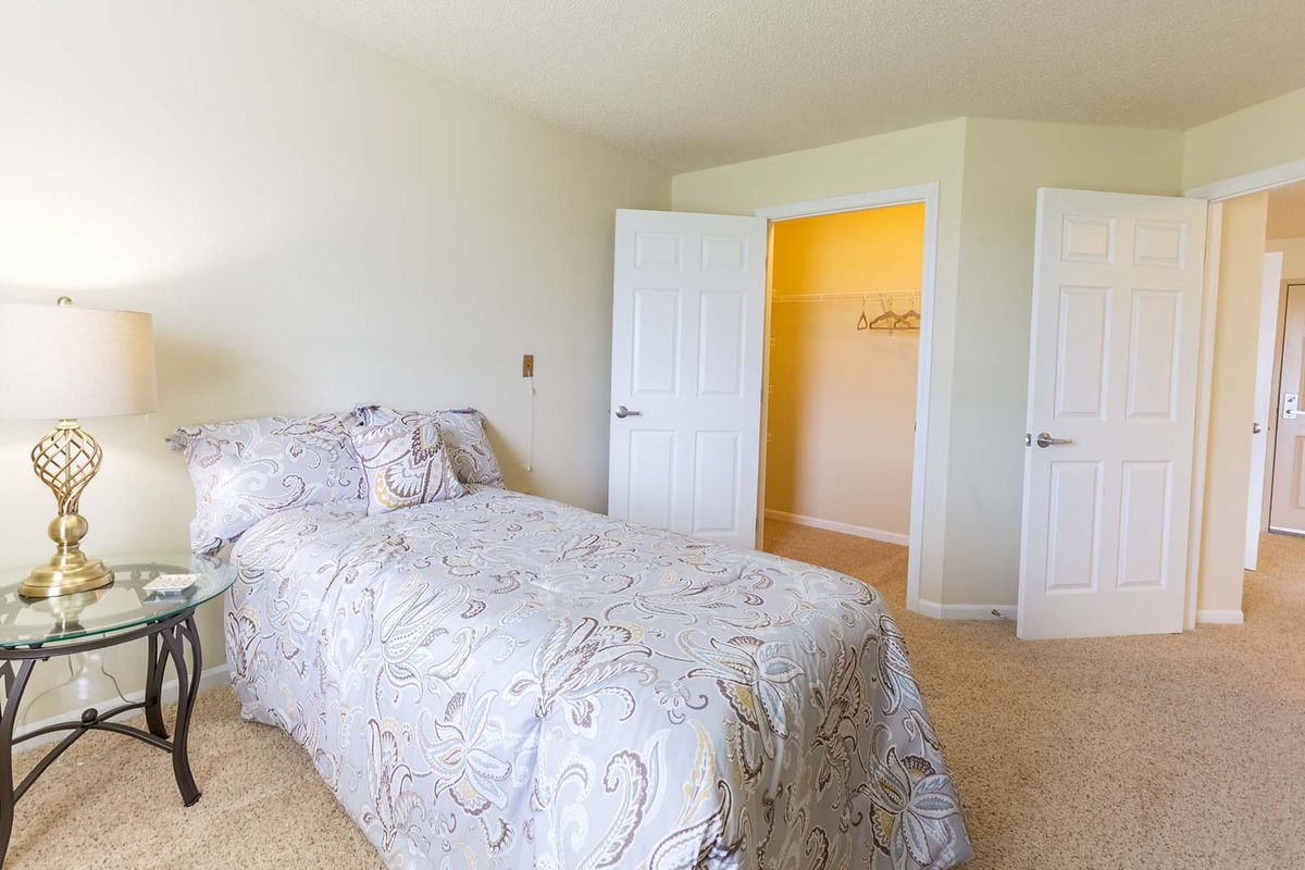A bright bedroom with a bed and open closet doors, featuring light-colored walls and carpet.