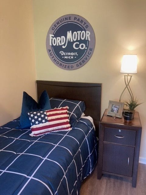 A cozy bedroom featuring a Ford Motor Co. sign, American flag pillow, and stylish nightstand with a lamp.