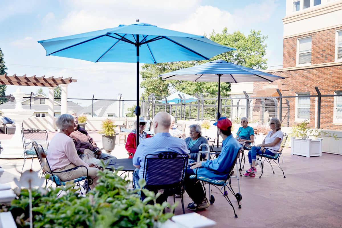 A group of people socializing under blue umbrellas on a sunny patio.