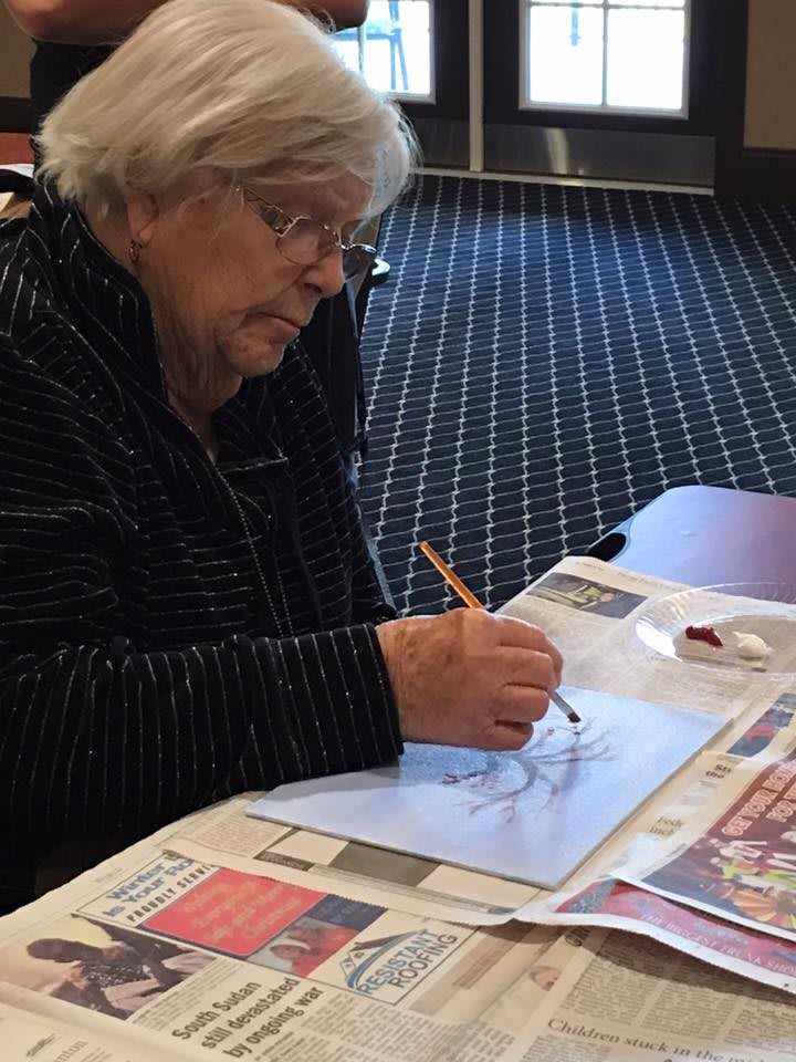 An elderly woman is drawing with a pencil on paper at a table covered with newspapers.