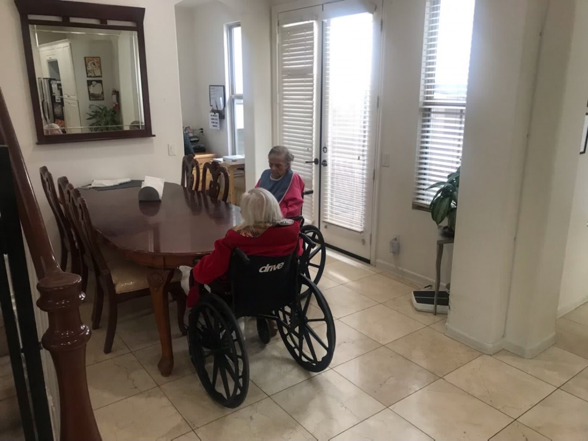 Two elderly individuals in wheelchairs sitting at a dining table in a bright, tiled-floor room.