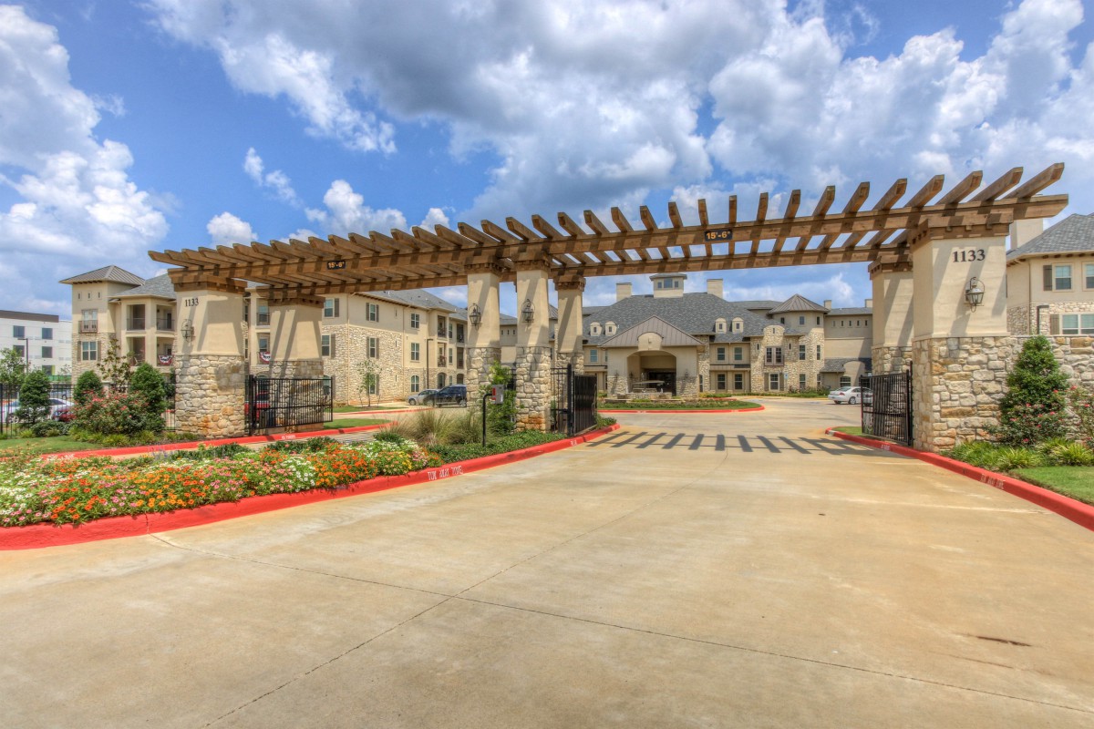 Entrance to a modern apartment complex featuring a pergola and landscaped surroundings under a cloudy sky.