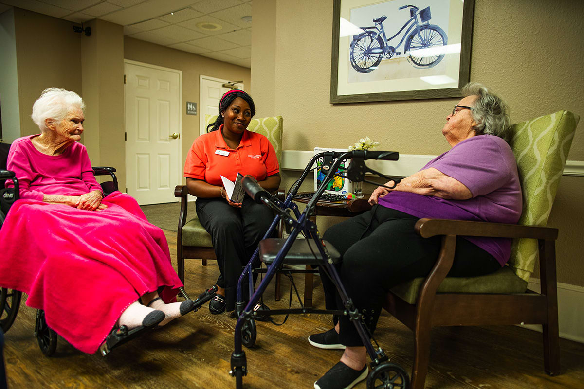 A caretaker sitting with two elderly women, one in a wheelchair and another with a walker, engaged in conversation.