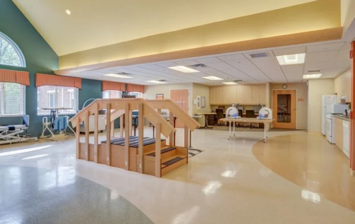 A spacious rehabilitation center therapy room featuring exercise equipment, a wooden staircase, and parallel bars.