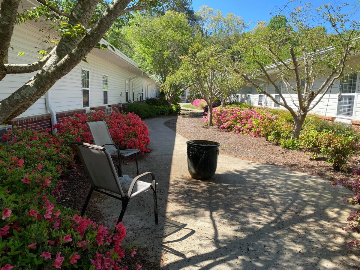 A peaceful garden walkway lined with colorful flowers and shaded by trees, featuring outdoor chairs for relaxation.