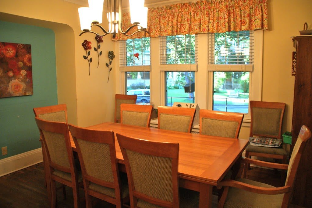 A cozy dining room with a wooden table and chairs, featuring large windows and a decorative light fixture.